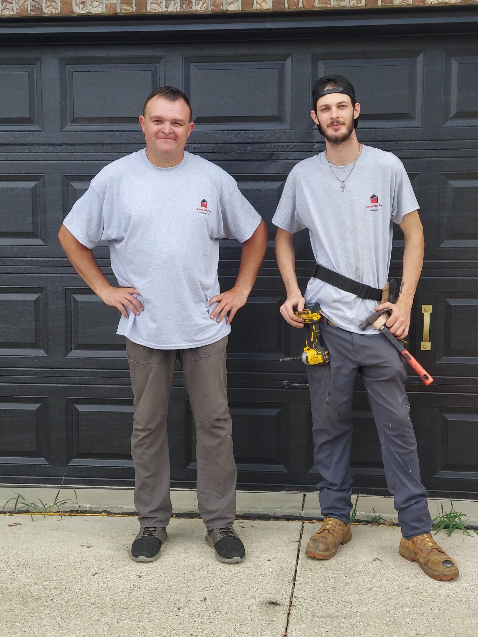 Two men in matching gray t-shirts standing in front of a garage door with tools.